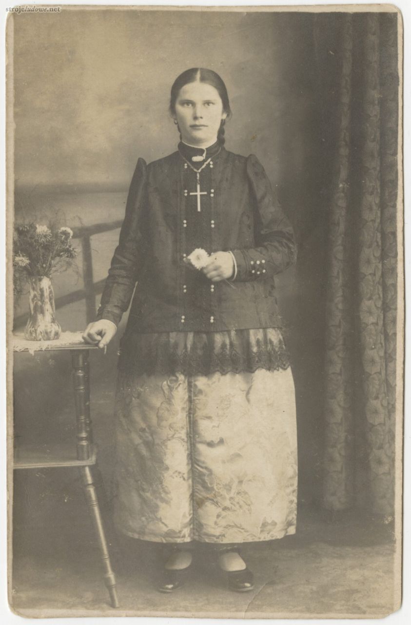 Portret młodej kobiety stroju pszczyńskim, na jakli symetrycznie ułożone dwa rzędy małych jasnych guziczków, na łańcuszku na jej szyi zawieszony jest duży krzyżyk, fotografia atelierowa, powiat pszczyński (miejscowość niezidentyfikowana – na rewersie zdjęcia brak pieczątki fotografa), lata 20-30. XX w., autor nieznany, z kolekcji Muzeum Śląskiego w Katowicach.