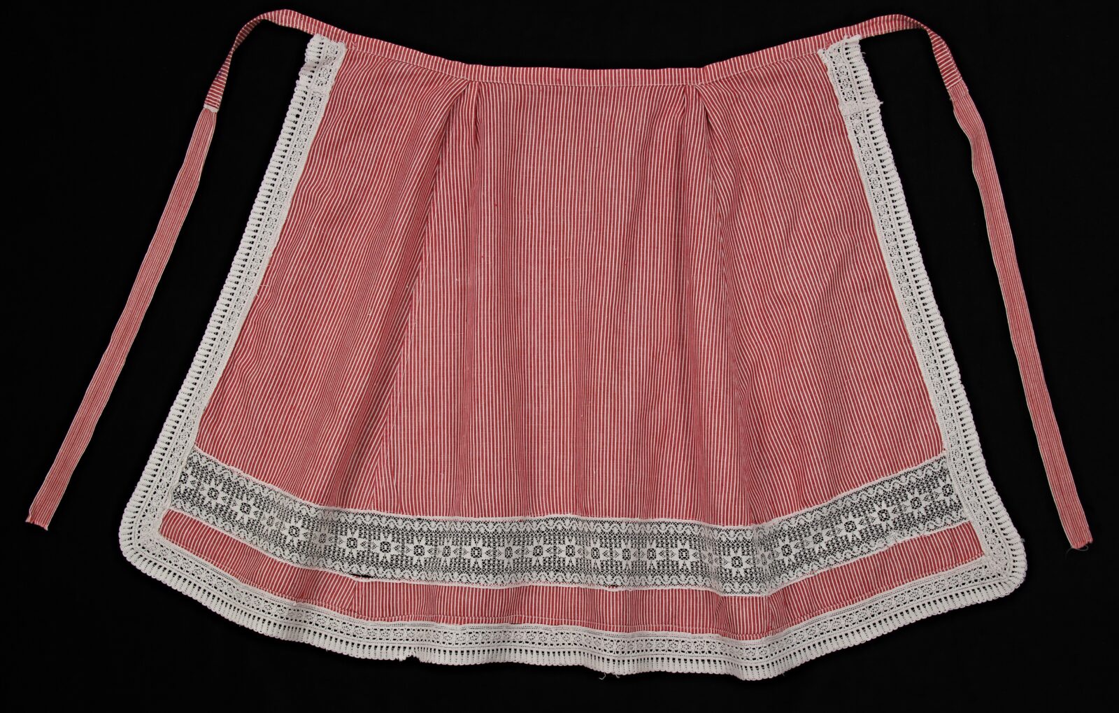 Fartuch uszyty z&nbsp;fabrycznego materiału bawełnianego, w&nbsp;biało-czerwone paseczki, wstawka i&nbsp;brzegi wykończone koronką fabryczną, wyk. nieznany, noszona do 1939&nbsp;r., Pałuki, zbiory Pałuckiej Izby Muzealnej w&nbsp;Kcyni, obecnie Gminne Centrum Kultury  i&nbsp;Biblioteki im. Klary Prillowej w&nbsp;Kcyni, fot. Marian Kosicki